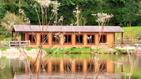 Lakeside view of a wooden cabin with a waterfront deck, surrounded by natural scenery.
